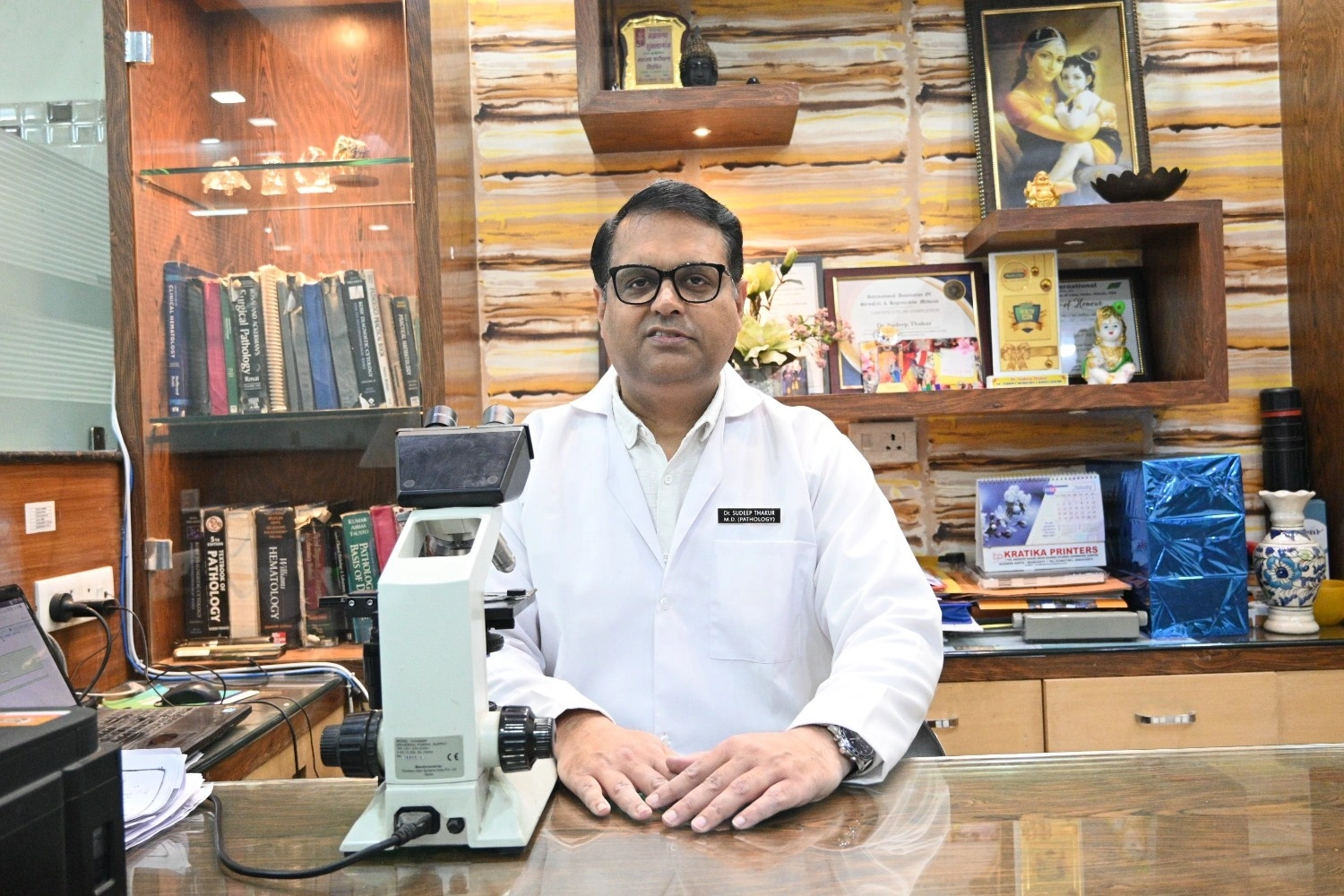 Dr. Sudeep Thakur, MD Pathology, at his desk with a clinical microscope and medical certifications.